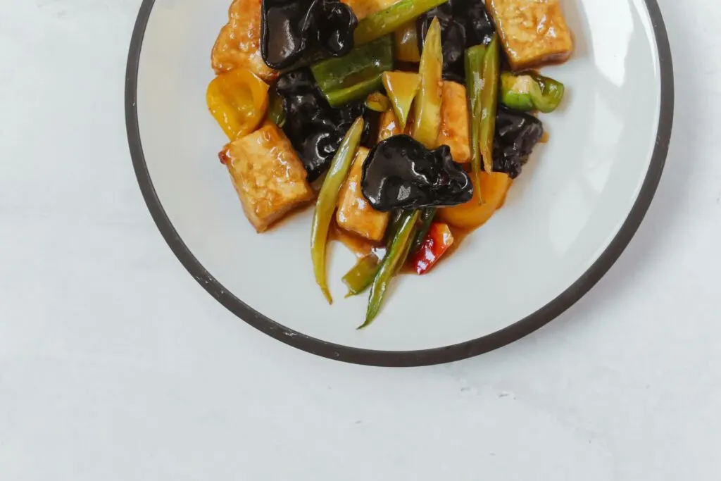 A plate of stir-fried vegetables and tofu with dark sauce, beautifully arranged on a light-colored surface.