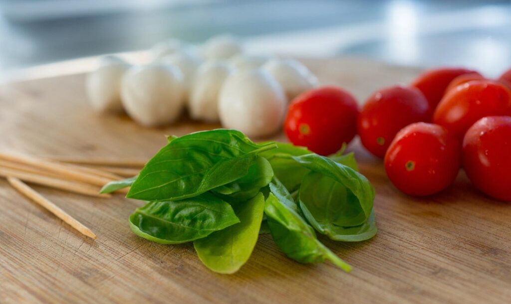 Fresh basil leaves, cherry tomatoes, mozzarella balls, and wooden skewers on a wooden cutting board, with a blurred background.