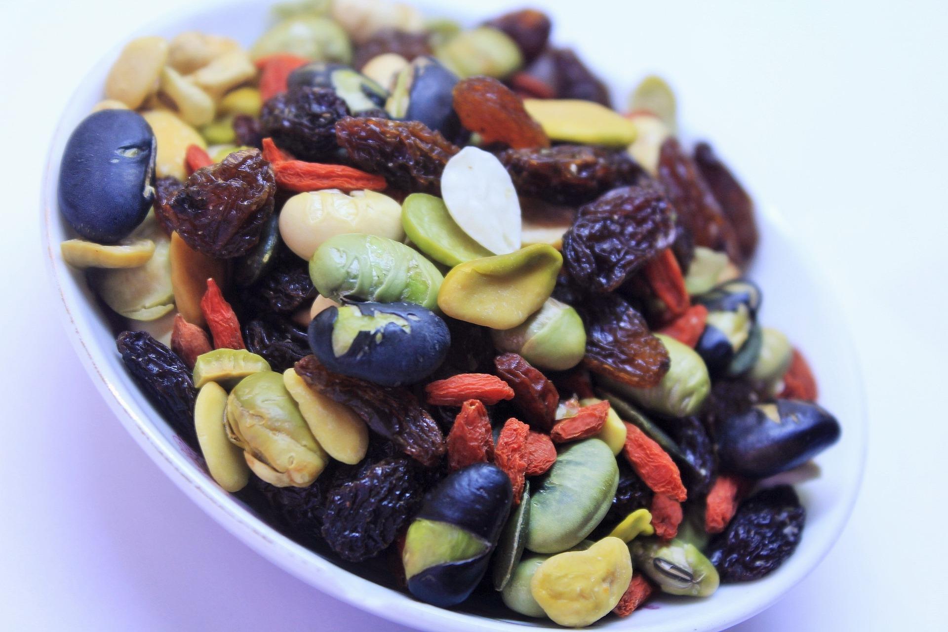 A close-up shot of a bowl filled with a variety of colorful dried fruits and nuts, including black, green, and red pieces, sitting on a white surface.