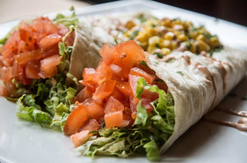 Close-up of two stuffed burritos on a plate, one filled with chopped tomatoes and shredded lettuce, and the other with corn kernels and drizzled with sauce.