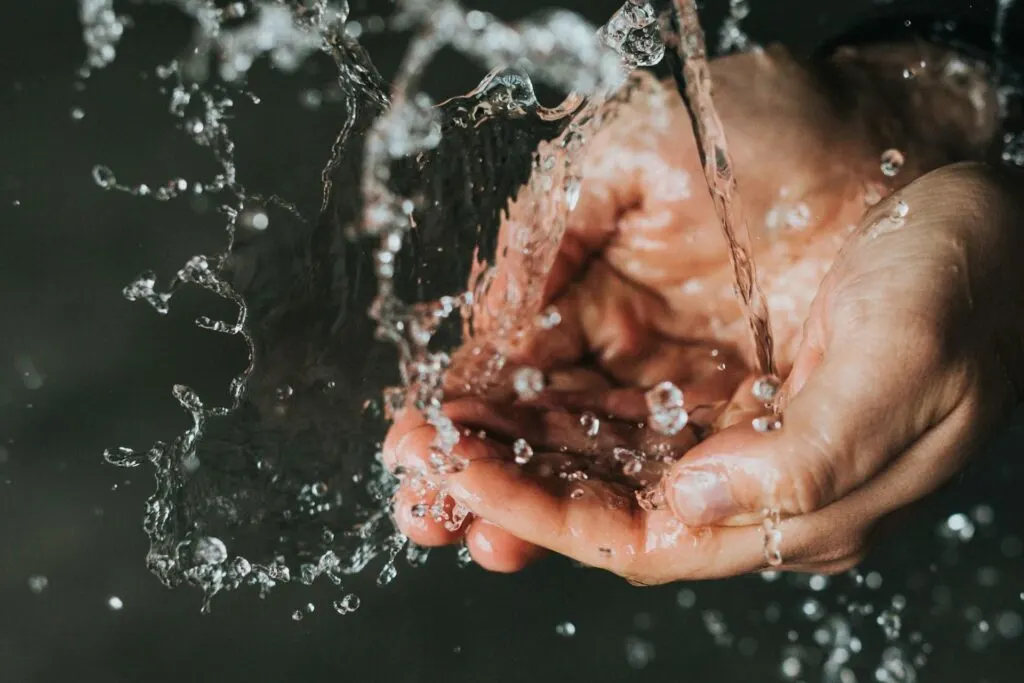 A close-up of water splashing over a pair of cupped hands, with droplets of water suspended in mid-air against a dark background.