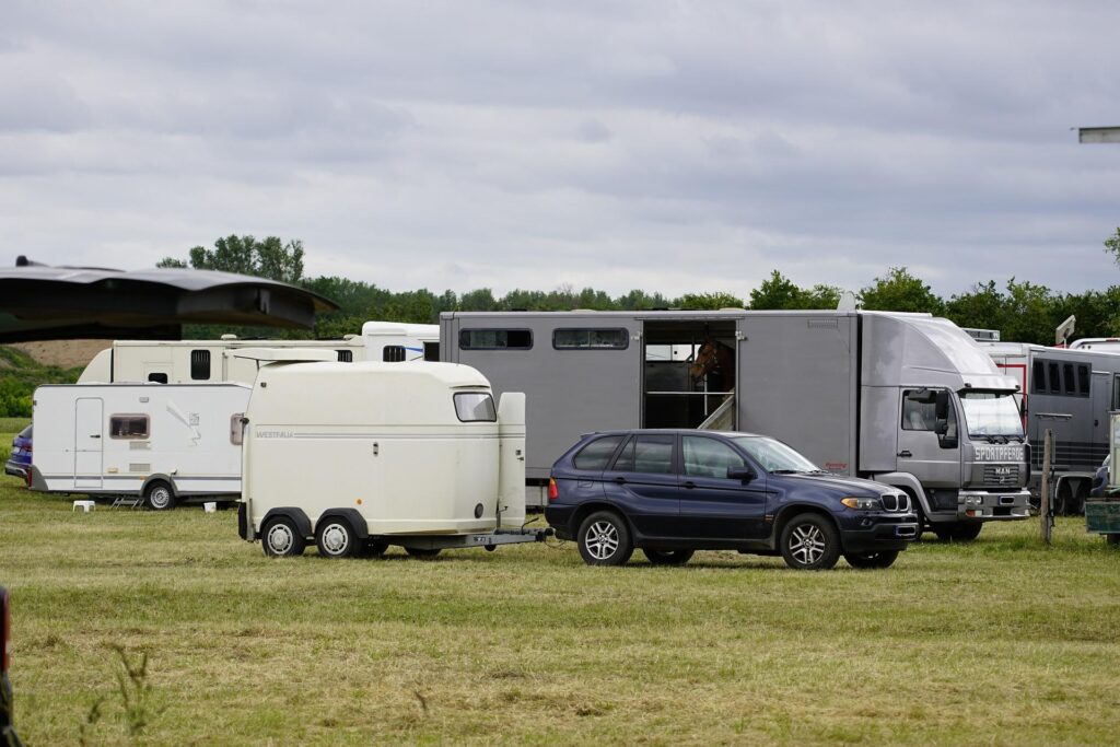 Various RVs and caravans parked in a grassy field under a cloudy sky. A navy blue SUV is seen towing a trailer, and there is a larger gray truck in the background.