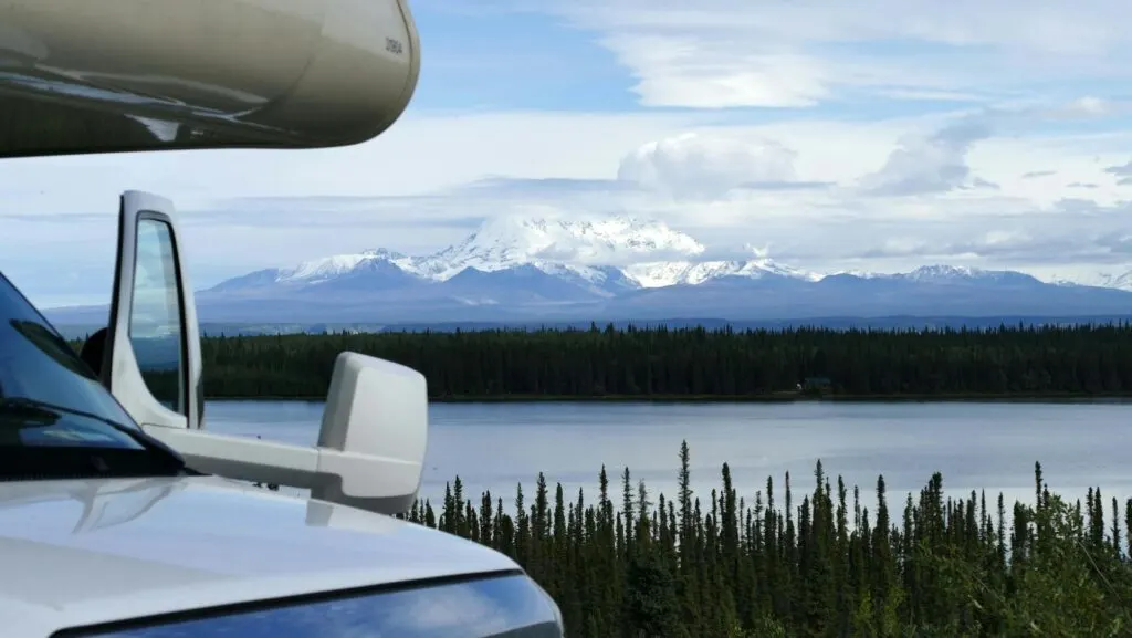 The side view of a camper van parked near a body of water, with a stunning mountain range covered in snow in the background.