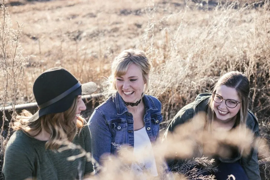 Three friends sitting in a dry, grassy field, chatting and laughing together.