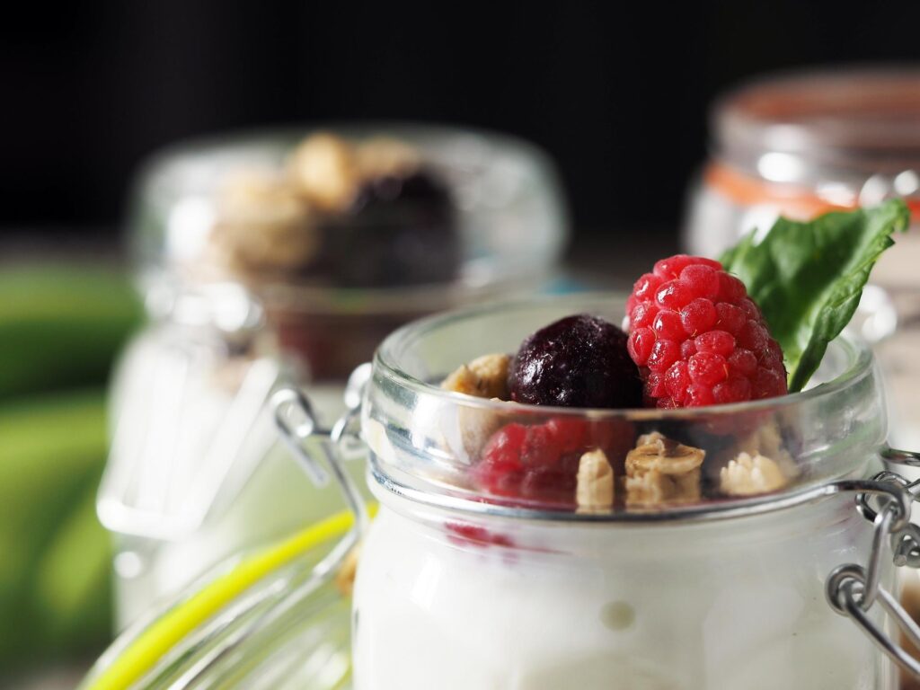 A close-up of a jar filled with yogurt, granola, a raspberry, and a blueberry, garnished with a mint leaf.