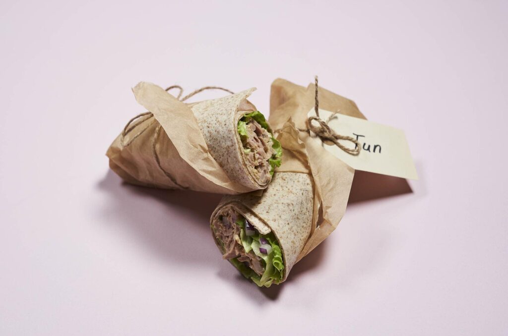 A pair of neatly wrapped sandwiches in brown paper with strings and a tag that reads "Tun," placed on a light pink surface.