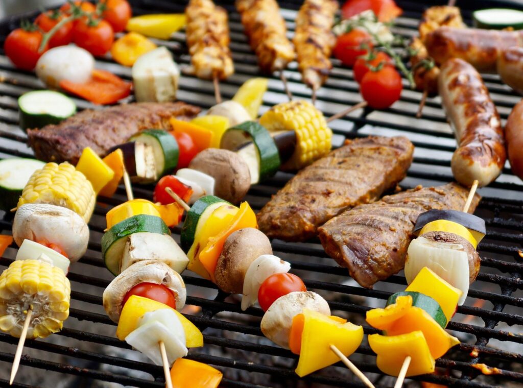 A grill loaded with a variety of foods, including vegetable skewers, corn on the cob, marinated meats, and sausages, all cooking over an open flame.