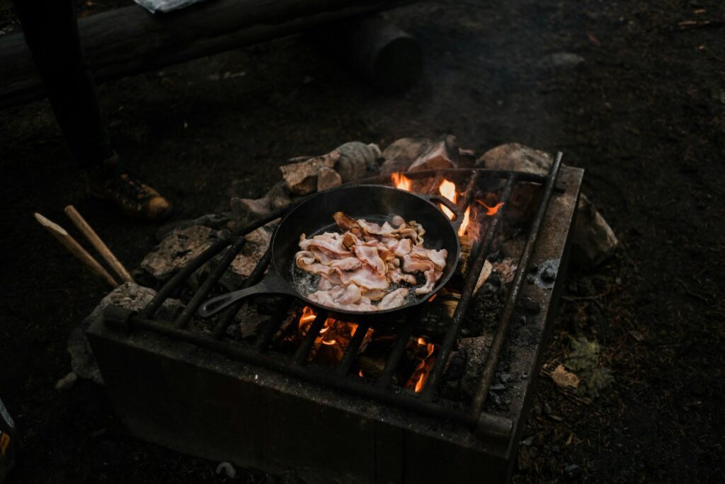 An outdoor campsite scene with a cast-iron skillet placed on a grill over an open flame. The skillet contains slices of bacon cooking, surrounded by rocks and logs. A person in hiking boots is partially visible in the background.