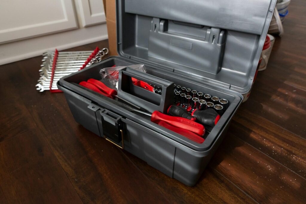 An open toolbox on a wooden floor, filled with various tools, including screwdrivers, sockets, and wrenches. Additional wrenches are laid out next to the toolbox.