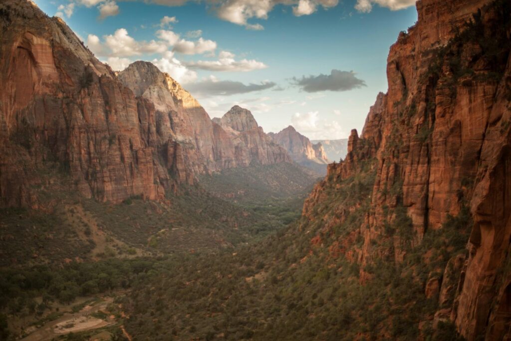 A dramatic canyon in Zion National Park with towering red rock cliffs and a green valley floor. The sky is partly cloudy, casting shadows and light over the rugged landscape.