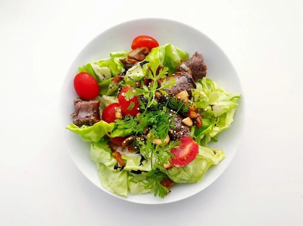 A white bowl of salad with lettuce, cherry tomatoes, meat, nuts, and topped with herbs.