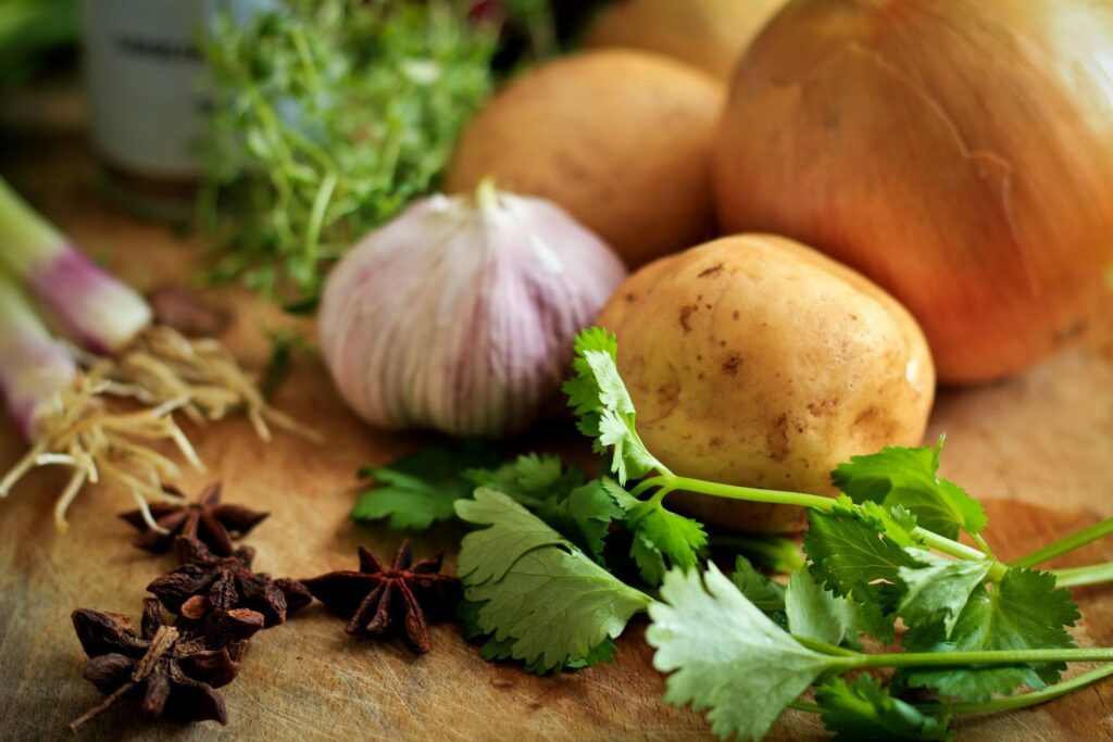 Various fresh vegetables and herbs, including garlic, an onion, potatoes, cilantro, star anise, and spring onions on a wooden surface.