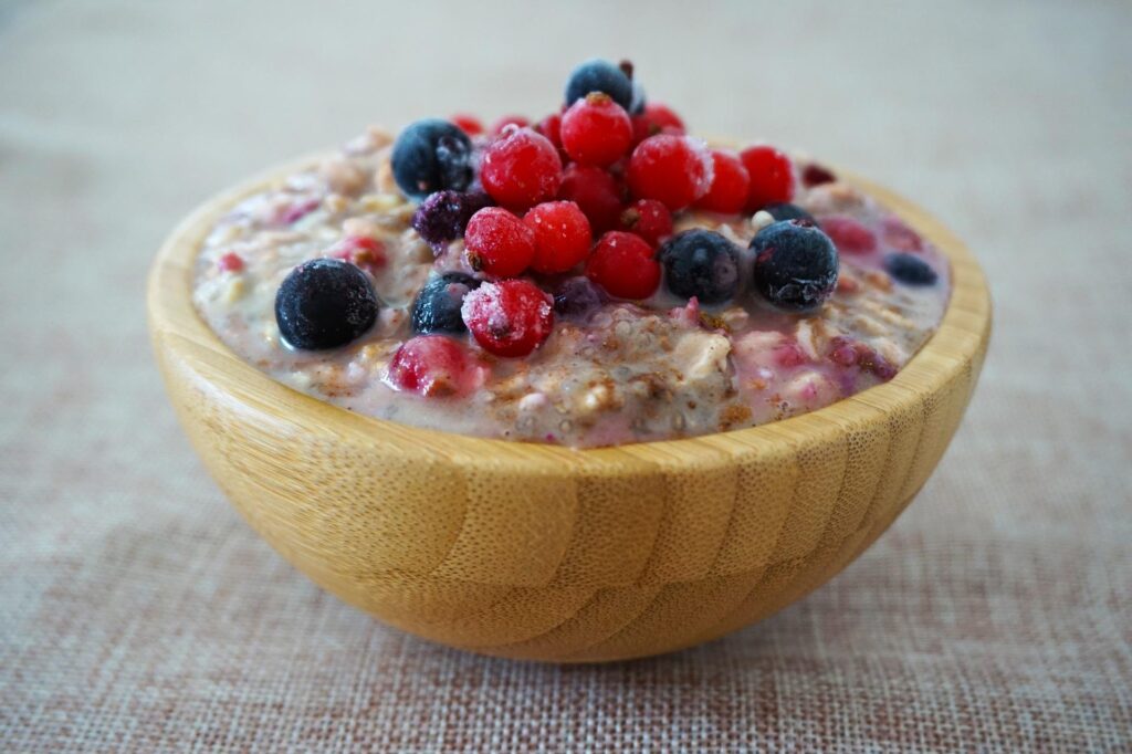 A wooden bowl filled with creamy oatmeal topped with a generous amount of fresh mixed berries, such as blueberries and red currants, on a beige tablecloth.