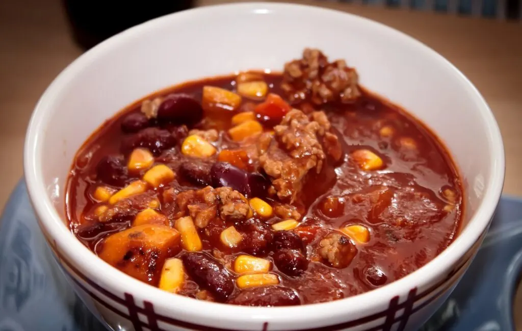 A bowl of chili with a rich tomato base, containing chunks of meat, kidney beans, corn, and other vegetables.