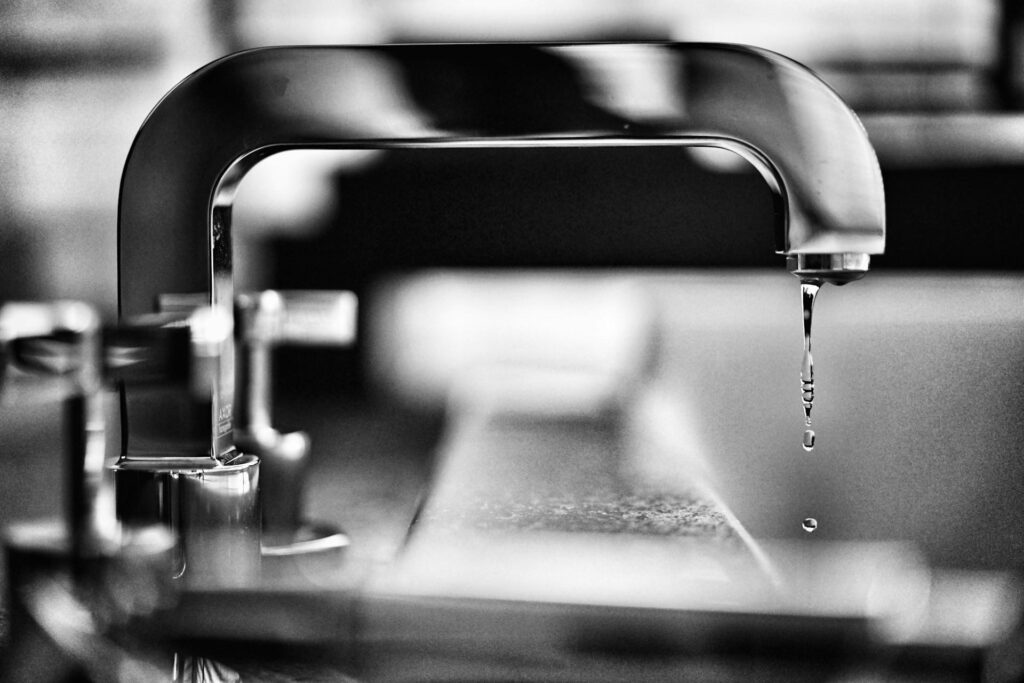 A black and white close-up view of a faucet with a single drop of water falling from the spout.