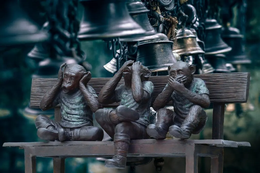 Three bronze monkey statues on a bench, representing the "See No Evil, Hear No Evil, Speak No Evil" proverb, with bells hanging in the background.