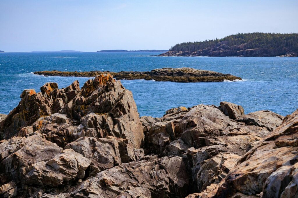 A coastal landscape in Acadia National Park with rugged rock formations and a vast expanse of blue ocean. The horizon shows distant islands and a clear blue sky.