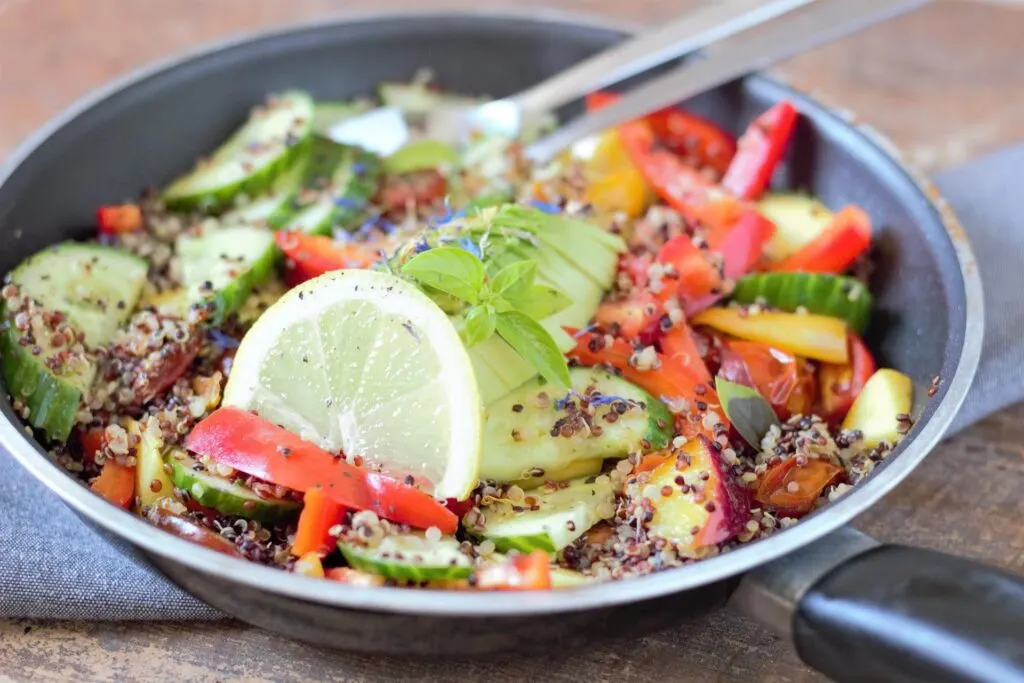 A pan of mixed vegetables and quinoa salad, garnished with lemon slice and basil, with a light rustic background.