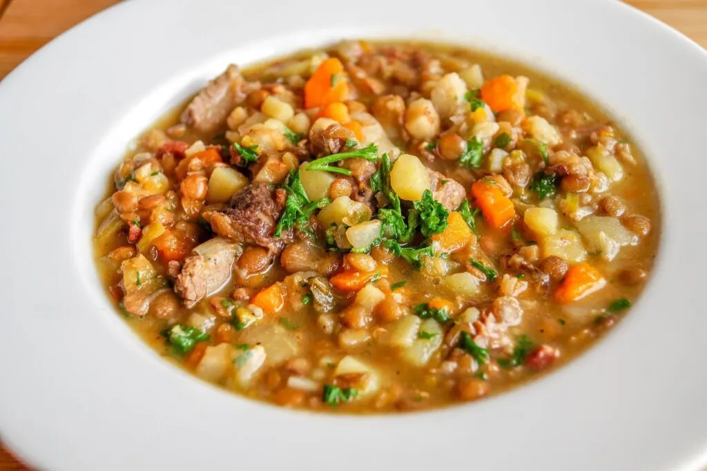 A bowl of hearty soup containing chunks of meat, carrots, potatoes, lentils, and garnished with fresh herbs, served in a white bowl.