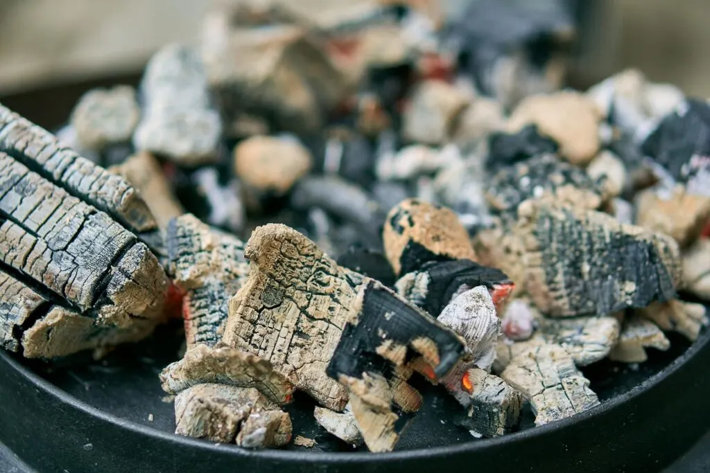 A close-up of charred pieces of wood and charcoal in a barbecue grill, with a mix of blackened and ashy textures.