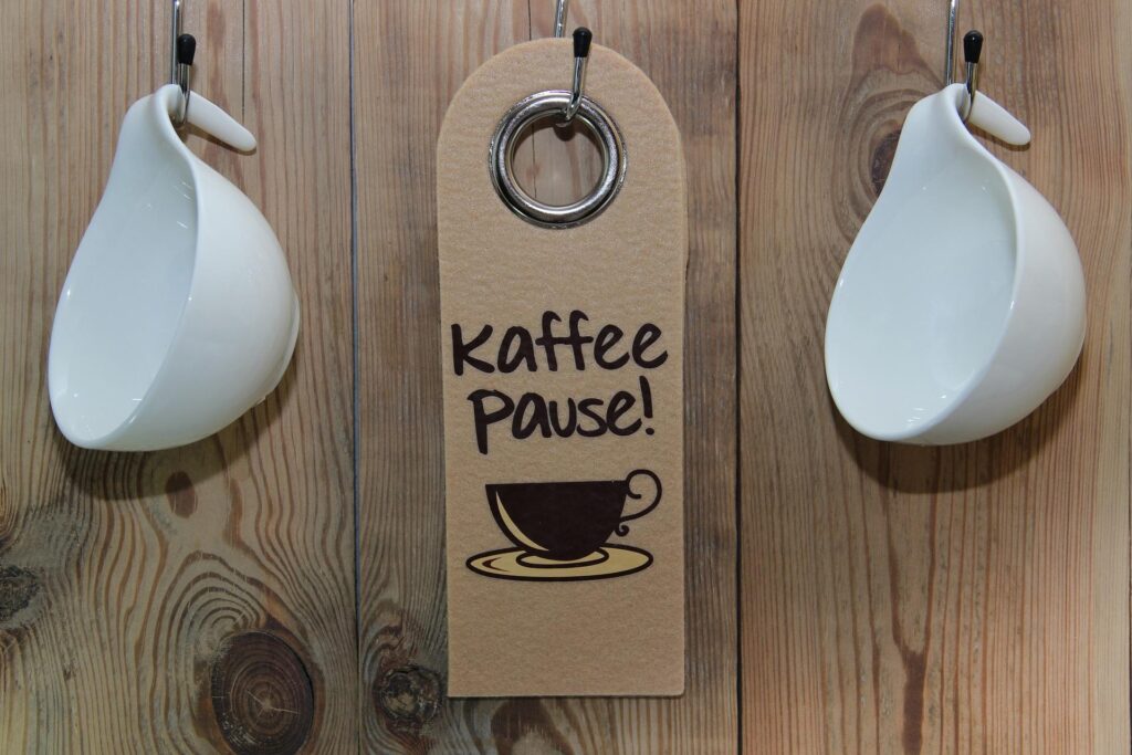 A wooden wall with two hanging white ceramic mugs flanking a sign that reads "Kaffee Pause!" with an image of a coffee cup and saucer.