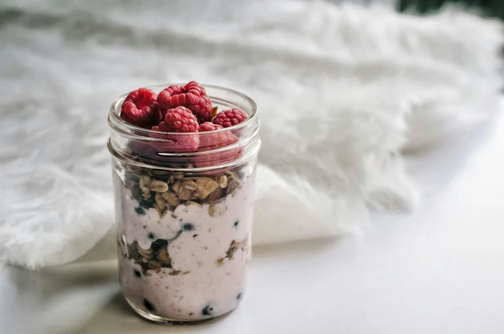 A jar filled with yogurt, granola, and raspberries, with a white, fluffy background.