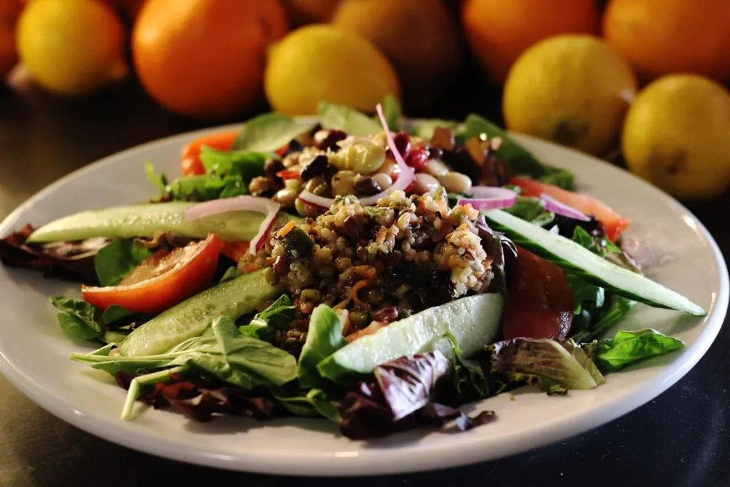 A plate of fresh salad with a variety of ingredients, including cucumber slices, tomato wedges, leafy greens, red onion slices, and a mixture of grains and beans, with lemons and oranges in the background.