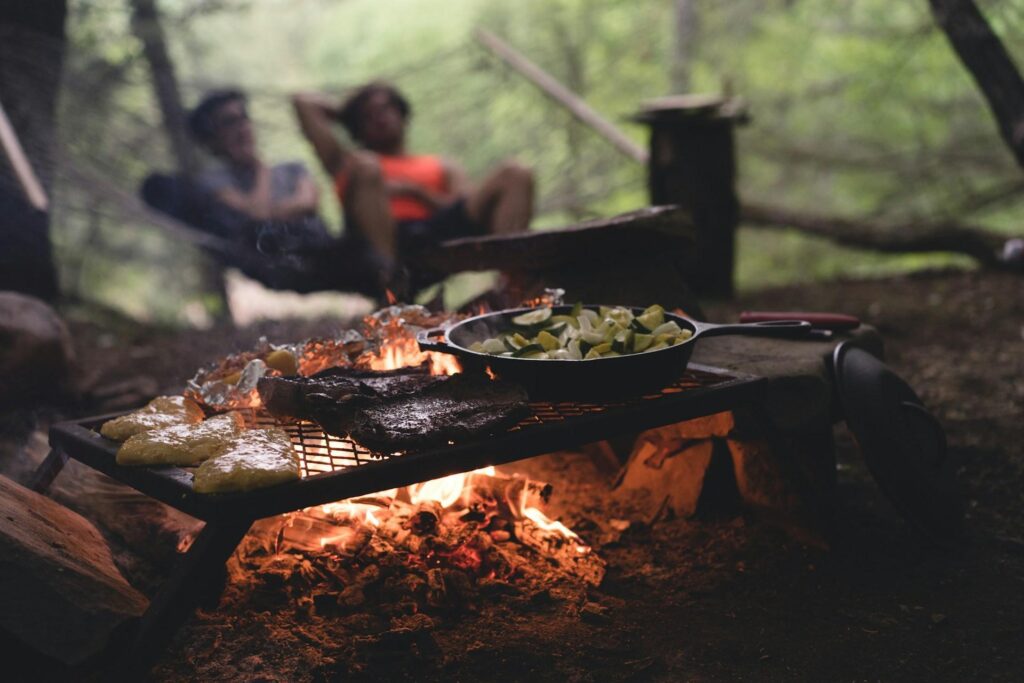 Outdoor campfire scene with a grill over an open flame, cooking meat and vegetables in a pan, with two people relaxing in a hammock in the blurred background surrounded by trees.