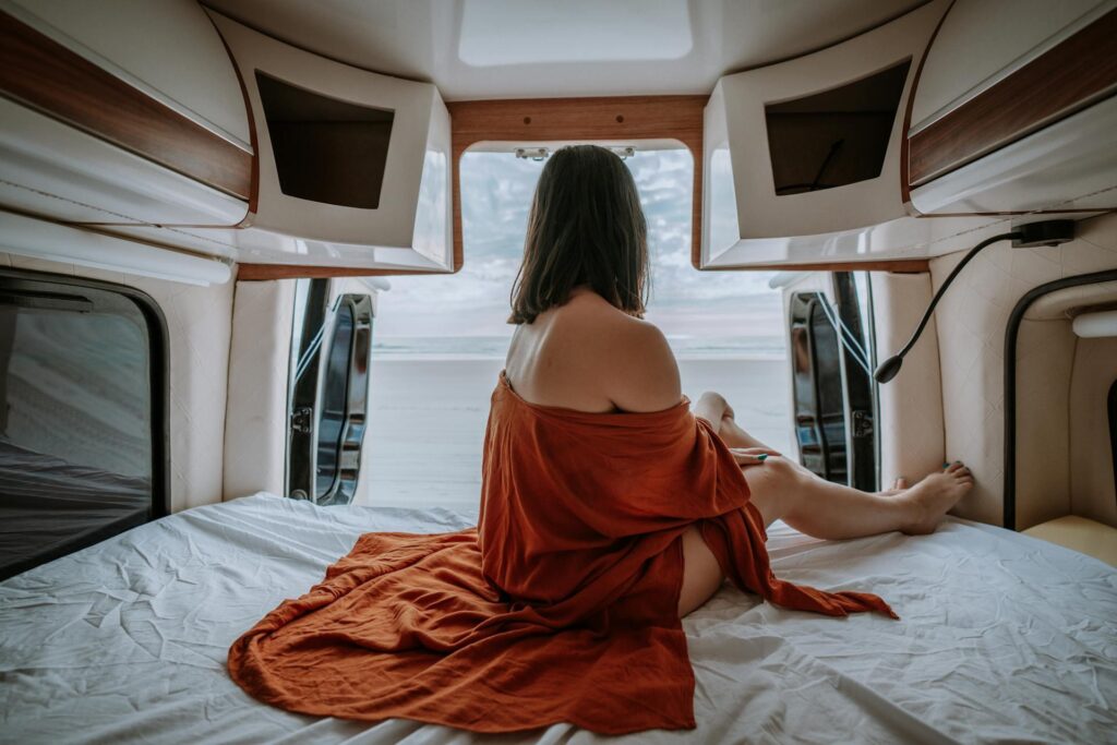 A cozy interior view of a camper van with a person sitting on a neatly made bed, facing an open door that reveals an outside view. The person is draped in an orange blanket, and the surrounding decor includes beige bedding and wooden storage compartments.