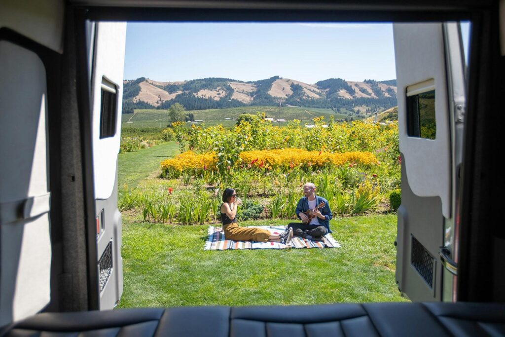 A view from inside a camper van looking out at two people sitting on a picnic blanket in a vibrant garden, surrounded by colorful flowers and green hills.