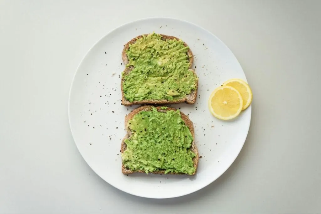 Two slices of avocado toast on whole grain bread, seasoned with black pepper, served on a white plate with two lemon slices on the side.