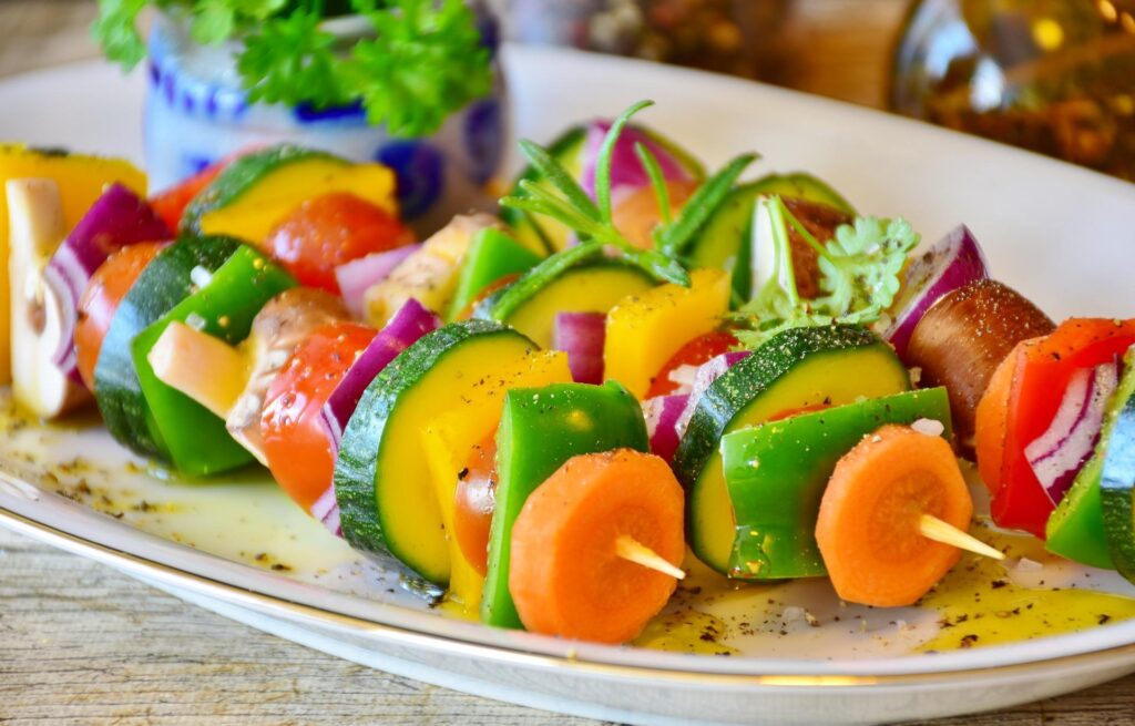 A plate of colorful vegetable skewers with pieces of green, yellow, and red bell peppers, zucchini, tomato, red onion, and carrot, garnished with herbs.