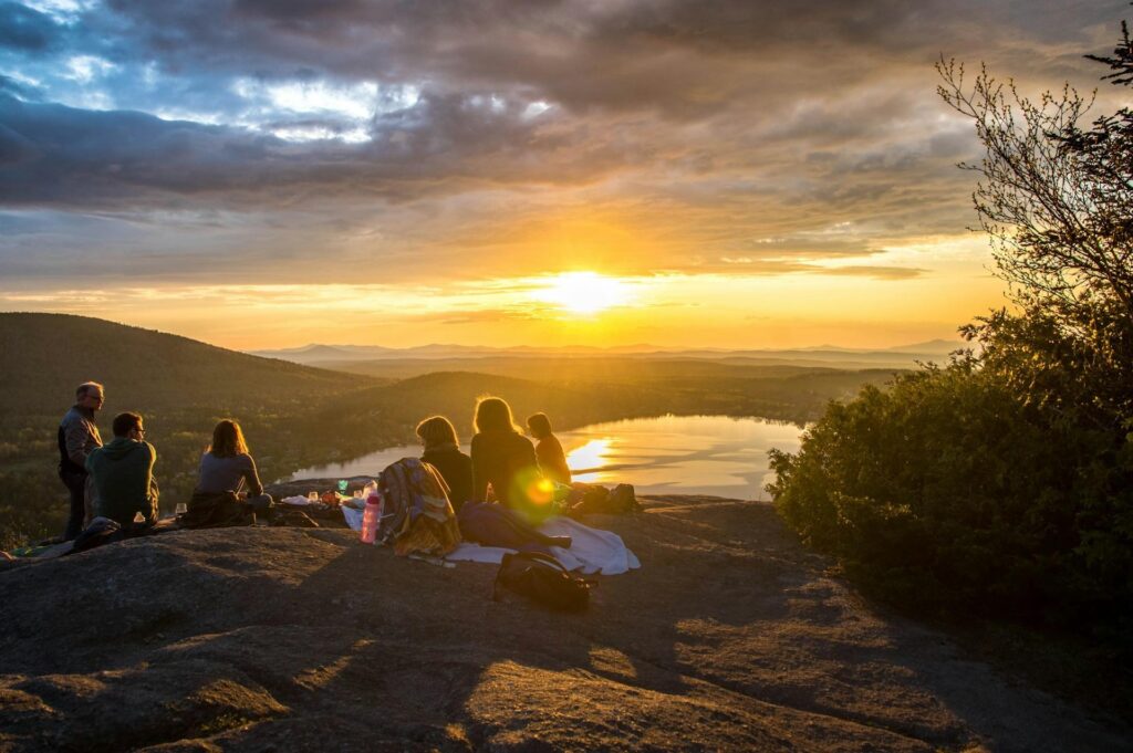 A group of people sitting on a rock enjoying the sunset view over a lake and mountains, with the sky painted in warm hues of orange and yellow.