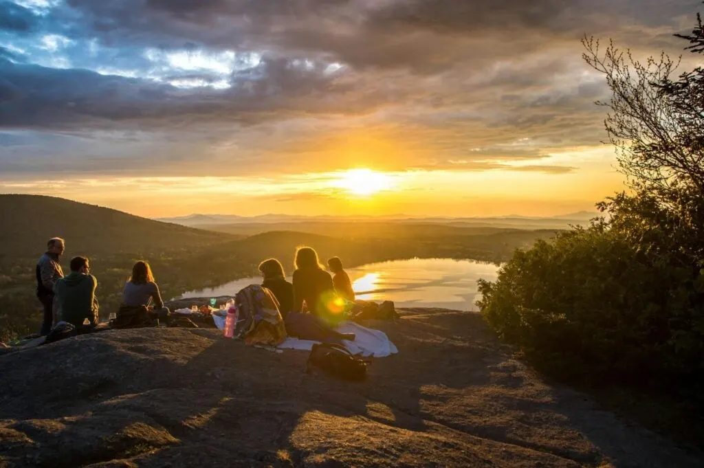 A group of people sitting on a rock enjoying the sunset view over a lake and mountains, with the sky painted in warm hues of orange and yellow.