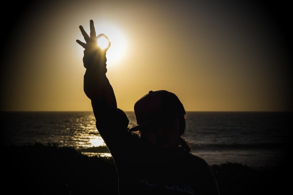 A silhouette of a person holding up their hand to the setting sun by the beach, creating a circular hand gesture around the sun.