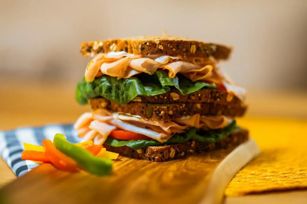 A stacked sandwich with whole grain bread, turkey, lettuce, and vegetables, with bell pepper slices on the side.