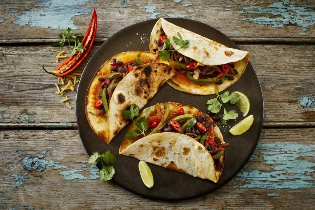 A top-down view of a plate with three stuffed tortillas filled with a mix of beans, peppers, onions, and garnished with cilantro and lime wedges, next to scattered cheese and a sliced chili pepper on a rustic wooden surface.