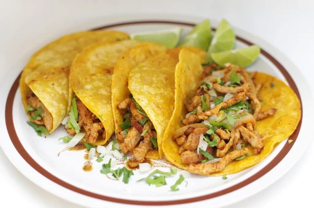 A plate of four yellow corn tacos filled with seasoned meat, diced onions, and cilantro, garnished with lime wedges on the side.