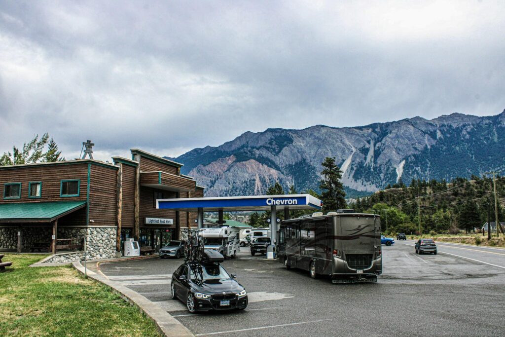 A Chevron gas station with multiple RVs and a car parked, set against a backdrop of mountains and a cloudy sky. The station includes a food mart and picnic tables on the side.