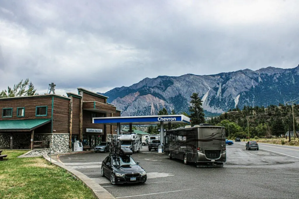 A Chevron gas station with multiple RVs and a car parked, set against a backdrop of mountains and a cloudy sky. The station includes a food mart and picnic tables on the side.