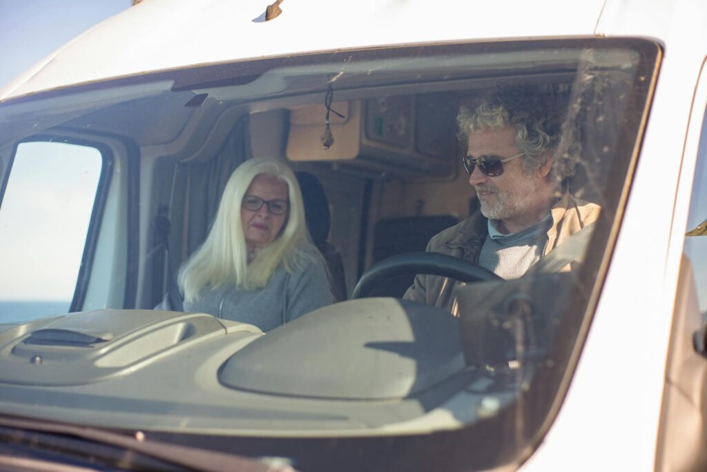 Two elderly people sitting together in the front seats of a vehicle, viewed through the windshield. Both are wearing glasses, and the driver is focused on the road while the passenger looks forward calmly.