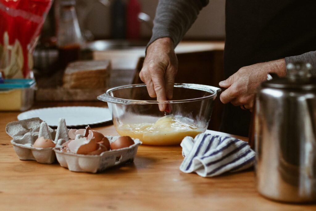 A person mixing ingredients in a glass bowl on a kitchen countertop, surrounded by eggshells and a striped towel.