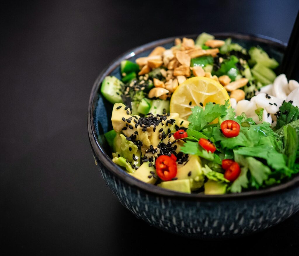 A bowl of mixed salad with avocado, cucumbers, greens, red chilies, lemon slice, and black sesame seeds.