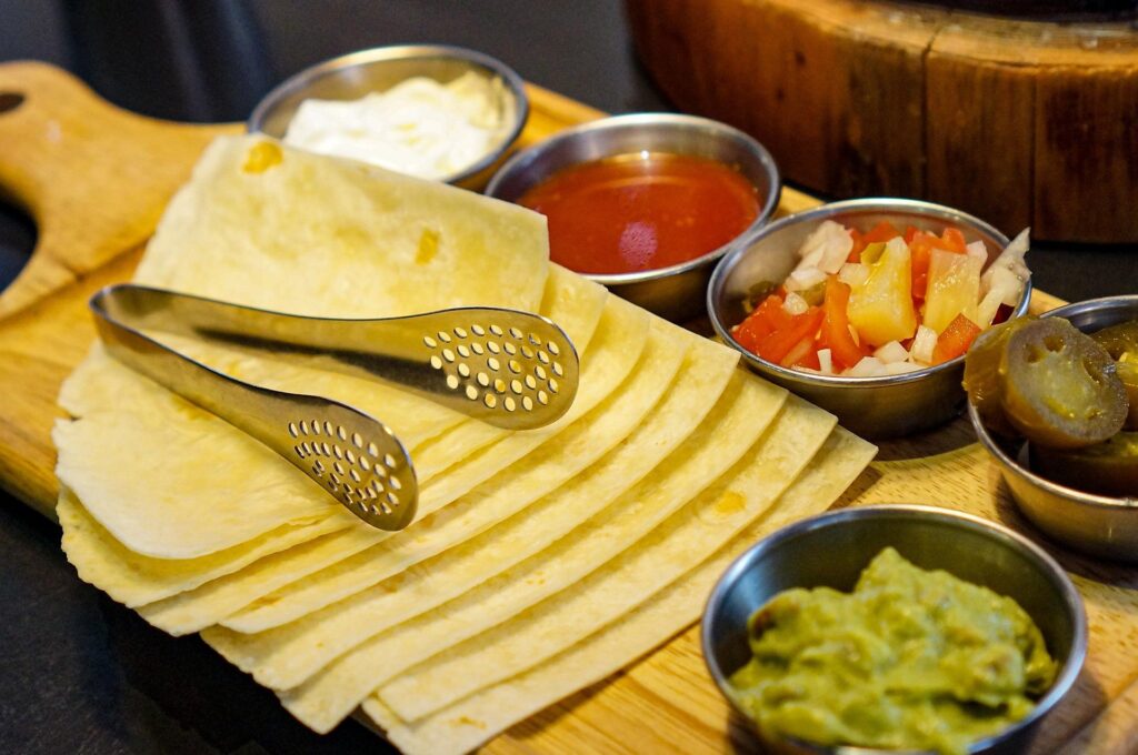 A wooden board with soft tortillas, metal tongs, and small bowls of various toppings including guacamole, sour cream, salsa, and jalapeños.