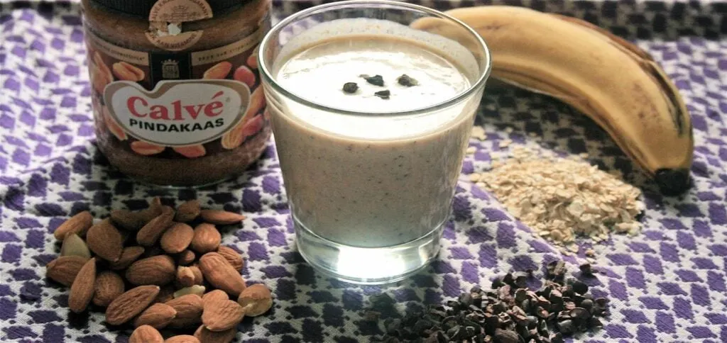 A glass of smoothie made with peanut butter and banana blend, surrounded by ingredients including a jar of peanut butter, banana, almonds, oats, and chocolate nibs, all placed on a patterned cloth.