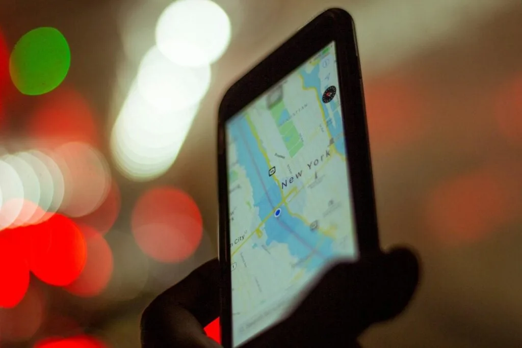 A hand holding a smartphone displaying a map with a location pinpointed on New York City. The background is blurred with bokeh lights in red and white.