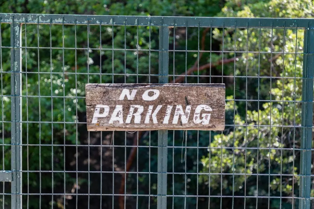 A rustic "No Parking" sign attached to a wire fence, with a background of green foliage.