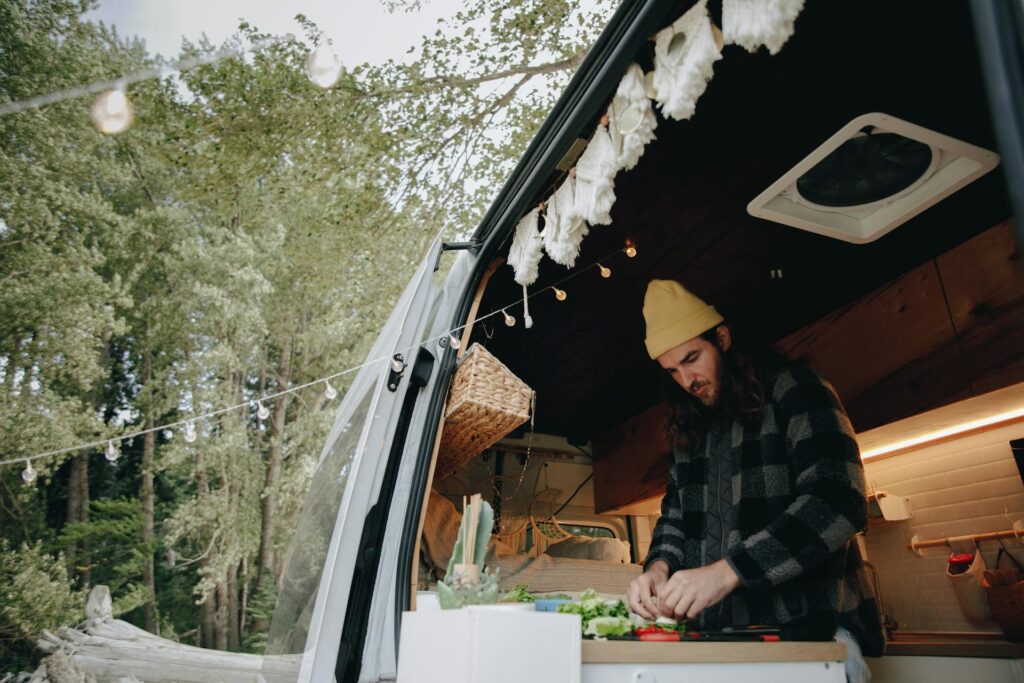 An open van kitchen with a person preparing food, decorated with string lights and surrounded by trees.