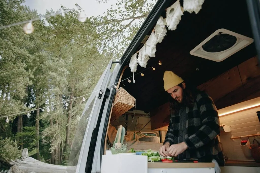 An open van kitchen with a person preparing food, decorated with string lights and surrounded by trees.