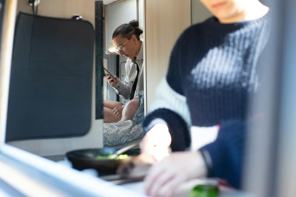 A candid photo of two people inside a small, cozy living space; one person in the foreground is chopping vegetables, while the other is sitting in the background, focused on their phone.